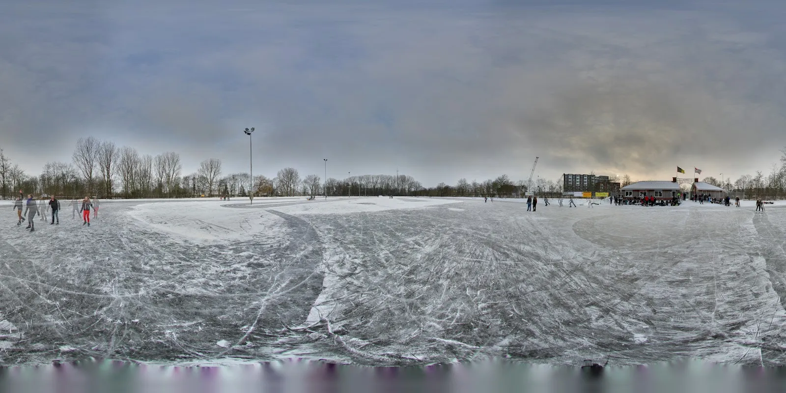 Schaatsers verspreid over het bekraste natuurijs van de Sneker ijsbaan in de winter, met de gebouwen en lichtmasten van de baan aan de horizon. – IJsbaan Dicky van der Werf (IJsclub Sneek)