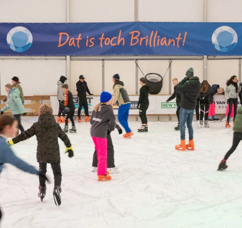 Children and adults skating across the white ice of the covered IJsbaan Duiven in front of a blue sponsor banner reading Dat is toch Brilliant — Brilliant IJsbaan Duiven