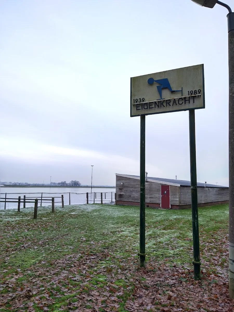 Het bord van Eigenkracht met de jaartallen 1939 en 1989 naast het lage houten clubgebouw en het onder water gezette baanveld aan de rand van Midwolda. – IJsbaan Eigen Kracht (Midwolda)