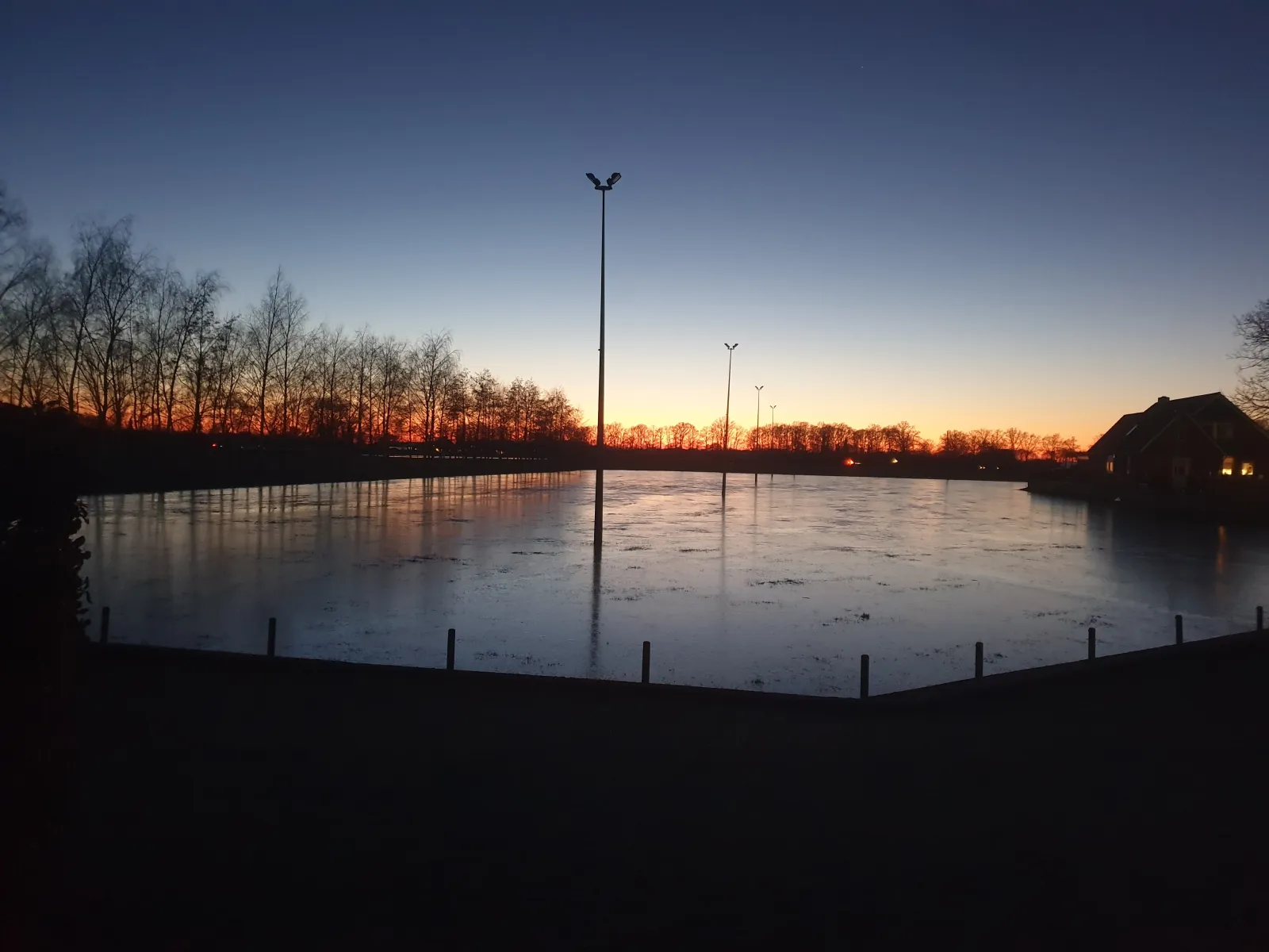 De ondergelopen weide als ijsbaan bij schemering, met lichtmasten langs de overkant en rechts een boerderij onder een oranje en blauwe avondlucht. – IJsbaan Lievelde