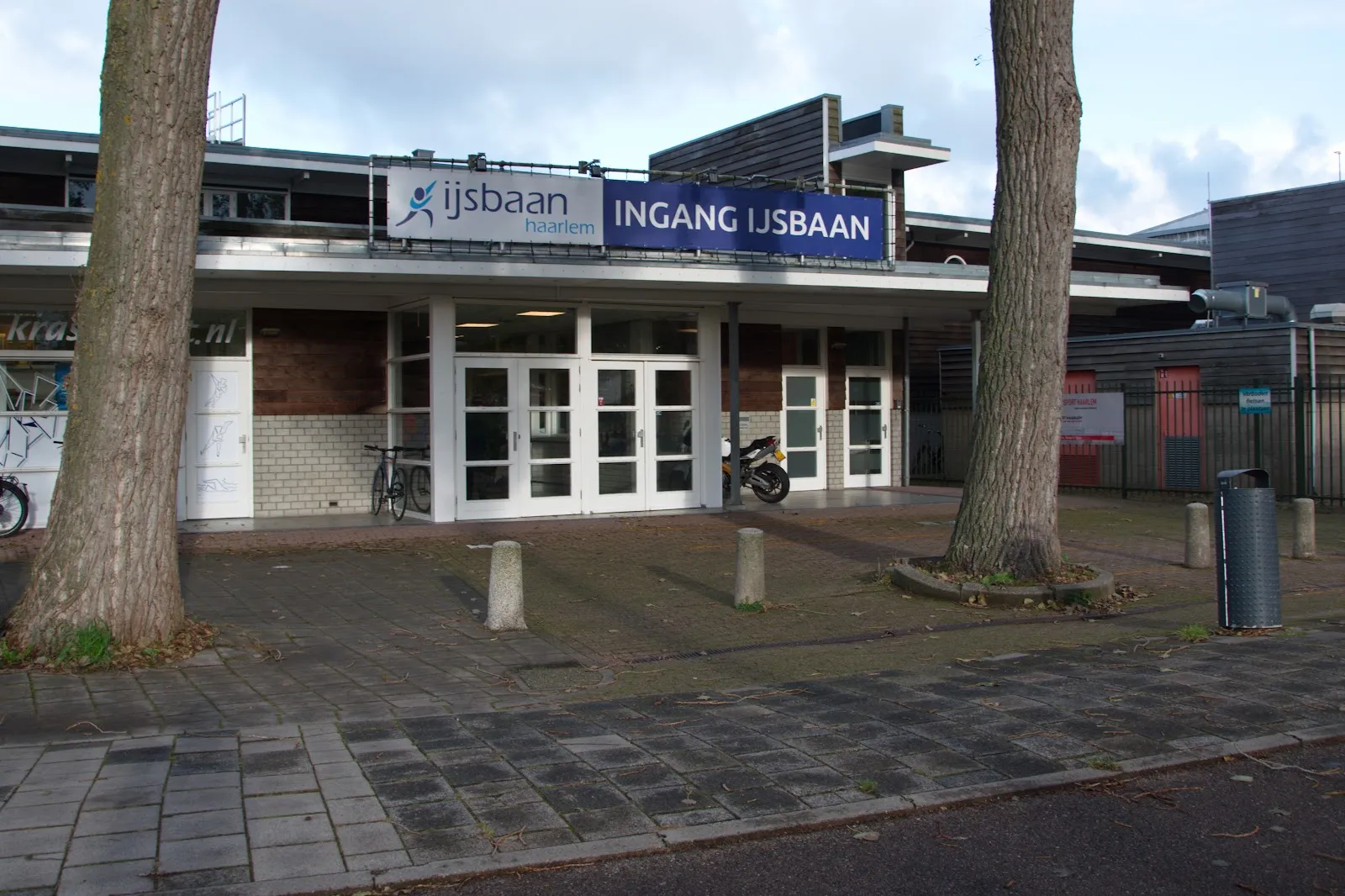 De ingang van de IJsbaan Haarlem onder het blauwe spandoek Ingang IJsbaan, met twee hoge bomen en de glazen deuren van het bakstenen gebouw – IJsbaan Haarlem