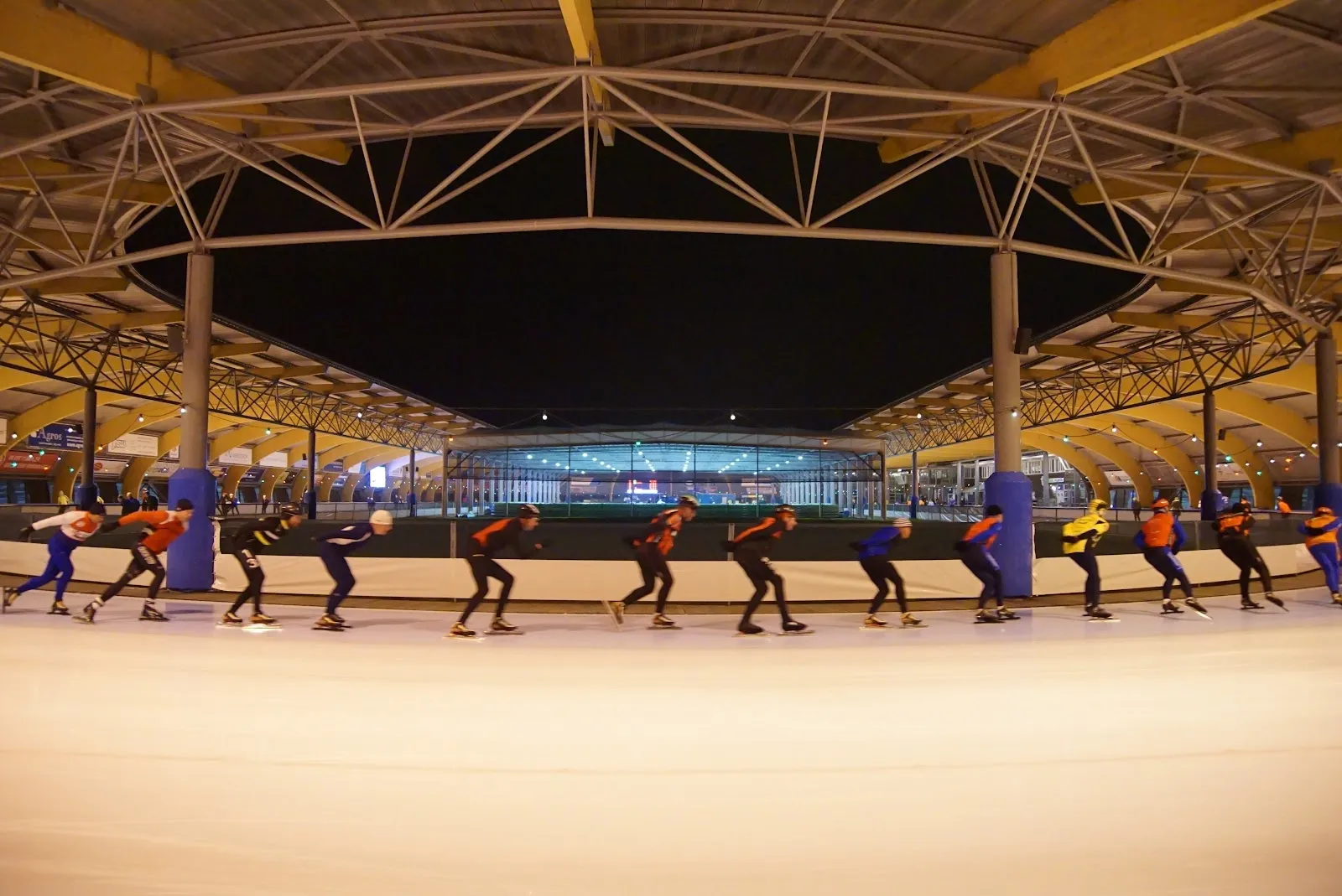 Schaatsers op de overdekte ijsbaan van IJsbaan Haarlem onder de geelhouten dakconstructie in de schemering. – IJsbaan Haarlem