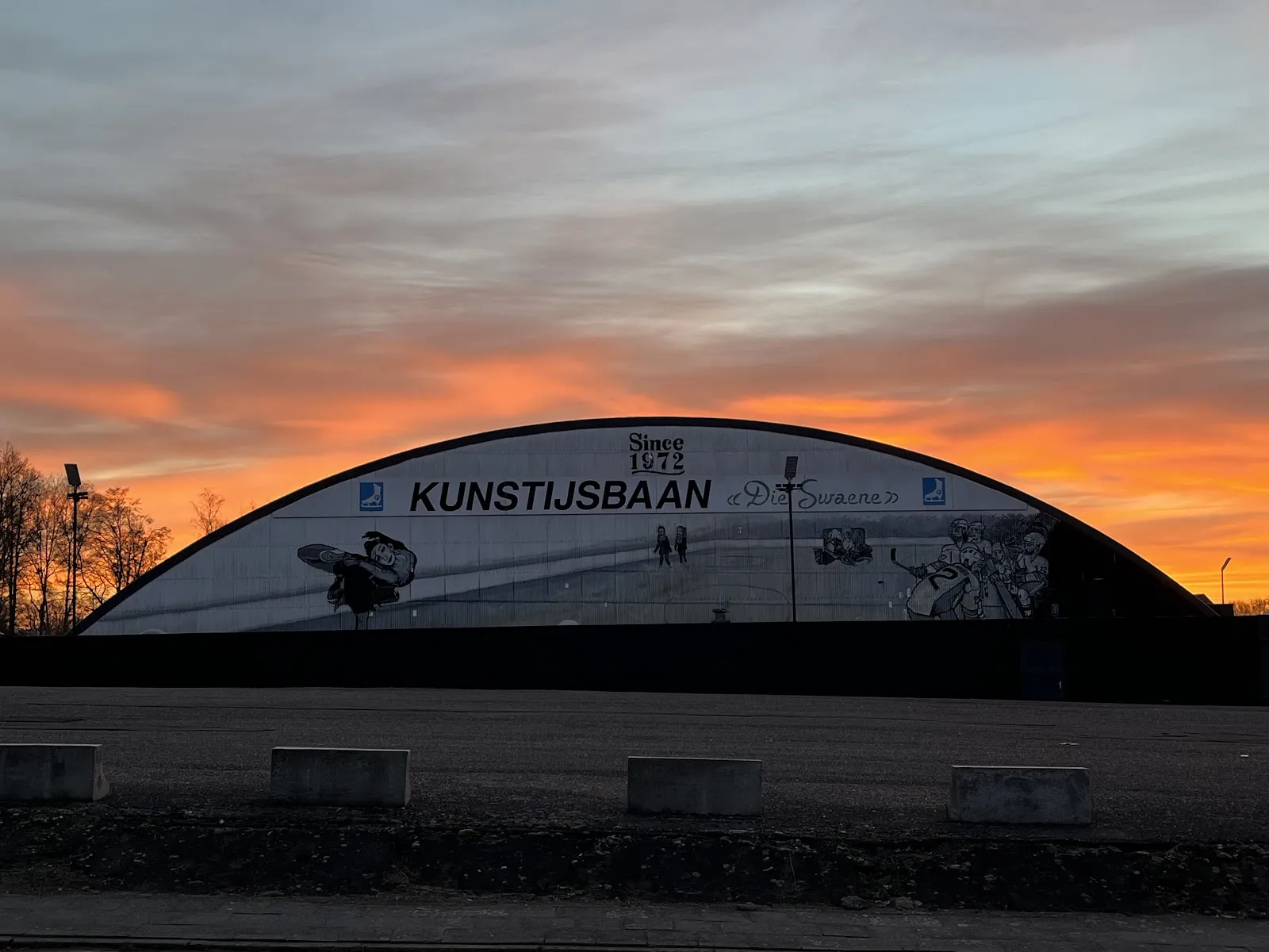 De gewelfde hal van IJsbaan Heist bij zonsondergang, beschilderd met “Kunstijsbaan, since 1972, Die Swaene” – IJsbaan Heist
