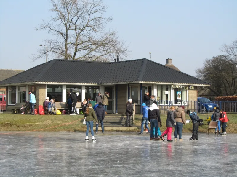Laag clubgebouw met een donker schuin dak naast het ijs, met bezoekers in de verte buiten. – IJsbaan Hoogenweg (HEO)