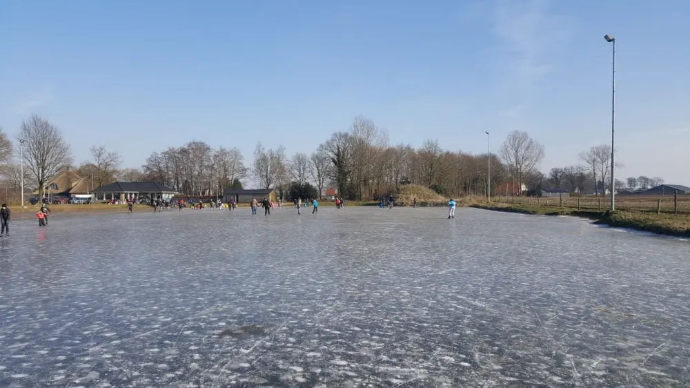 Wijds uitzicht over de bevroren baan van Hoogenweg met schaatsers in de verte en bomen langs de overkant. – IJsbaan Hoogenweg (HEO)