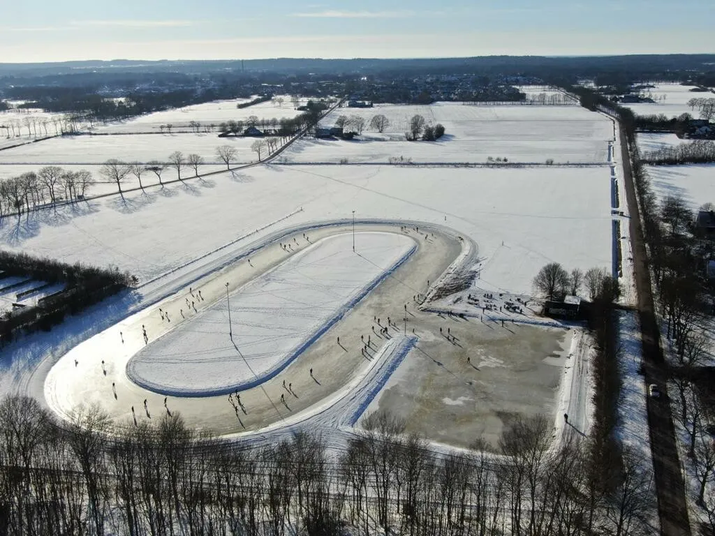 Aerial view of the Hennie Koersbaan in winter, with skaters spread along the oval ice track and its snow-covered infield among white fields. — Hennie Koersbaan (IJsclub Hulsen)