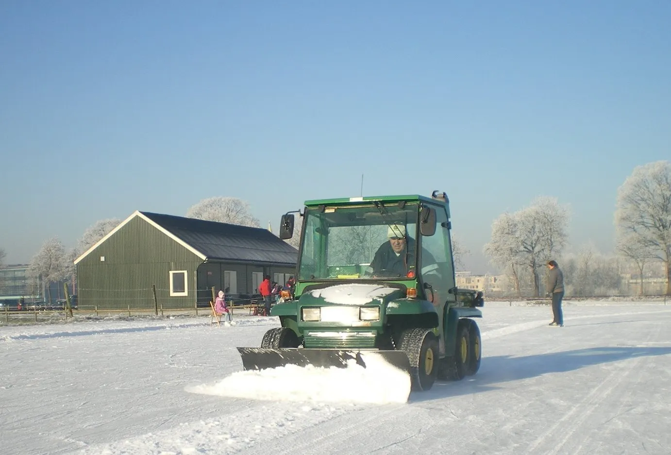 The dark green clubhouse and a green sweeper vehicle on the frosted HWZ rink under a clear winter sky. — IJsbaan HWZ