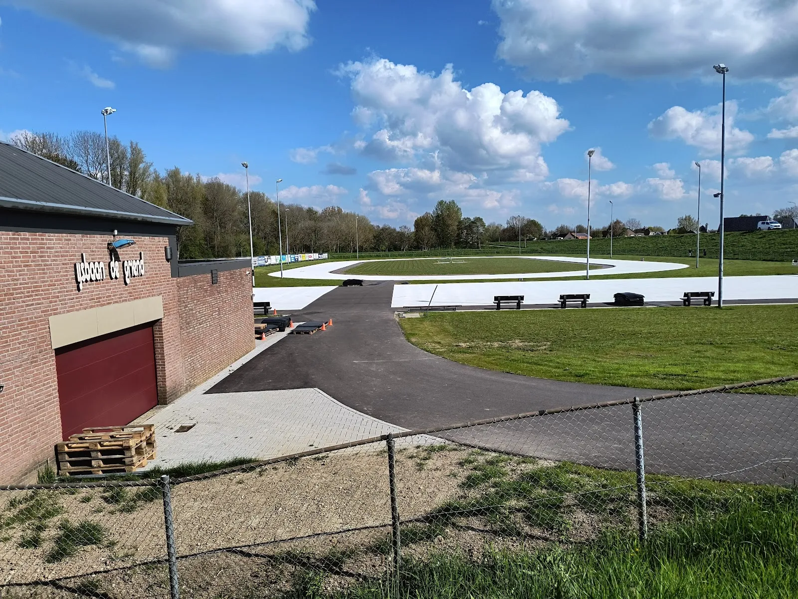 The brick clubhouse with the ijsbaan de griend sign beside the new pale track and floodlight masts under a bright spring sky. — IJsbaan Kerkdriel (de Griend)