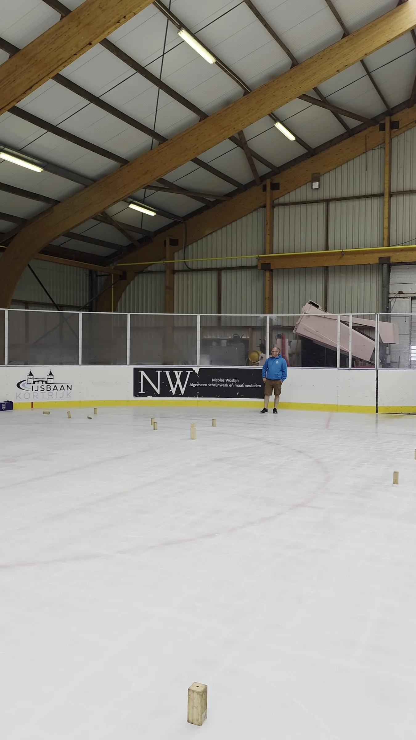 The ice at IJsbaan Kortrijk under the timber roof beams, with a few wooden kubb blocks on the ice and one person far along the boards — IJsbaan Kortrijk