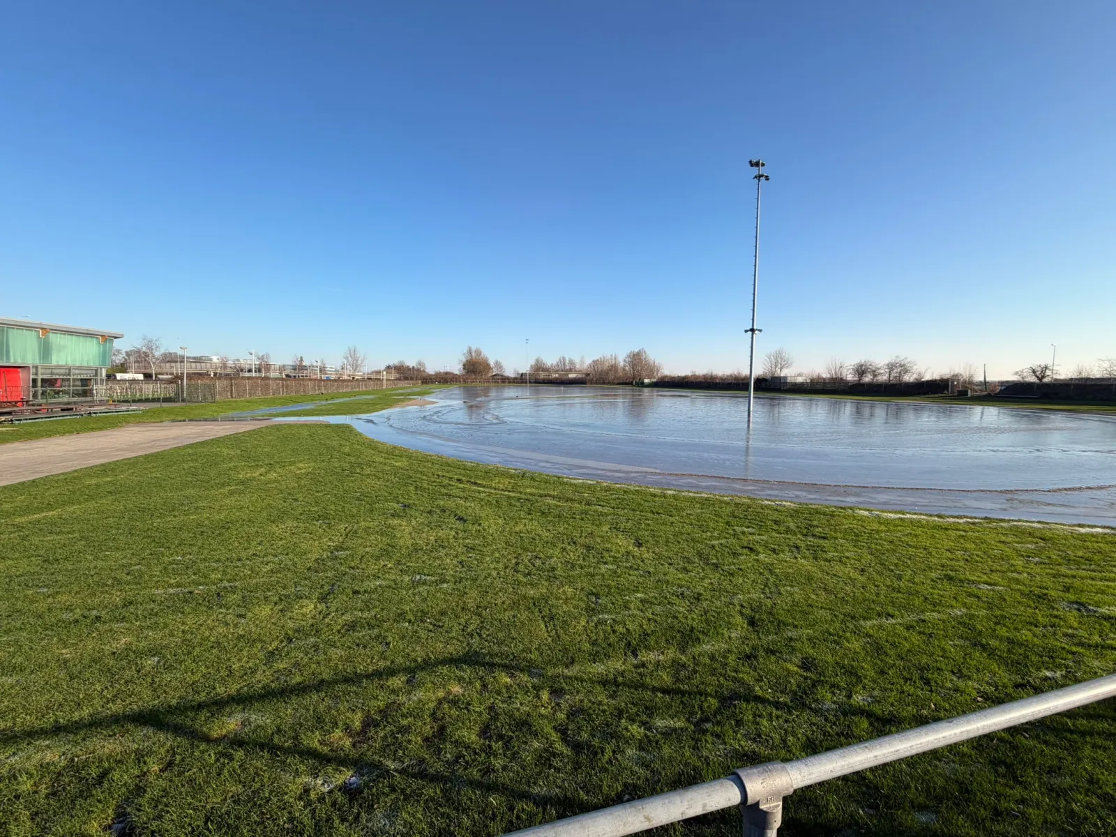 The flooded land rink in the Weteringpark under a clear blue sky, with a floodlight mast and the clubhouse roofs at the far edge of the oval. — Landijsbaan Weteringpark (Leiderdorp)