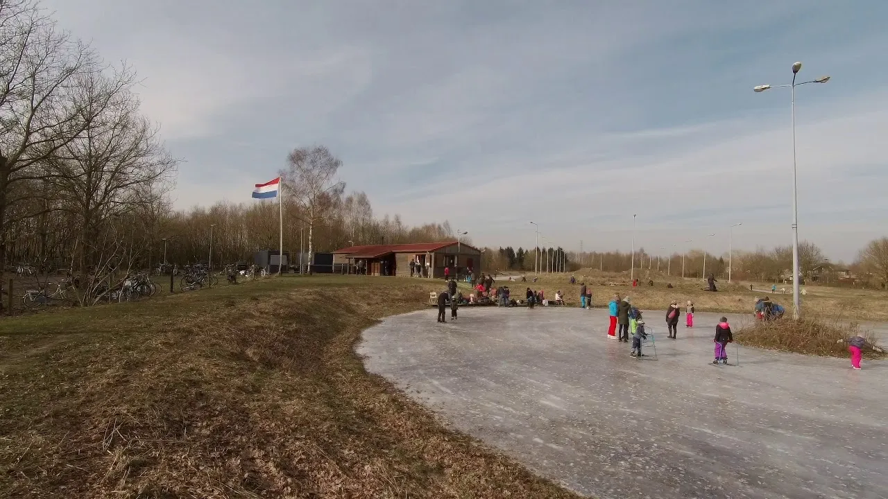 Skaters on the ice patch of the Lochem rink at Sportpark De Elze, with the red-roofed clubhouse, a flagpole and a floodlight mast behind. — IJsbaan Lochem