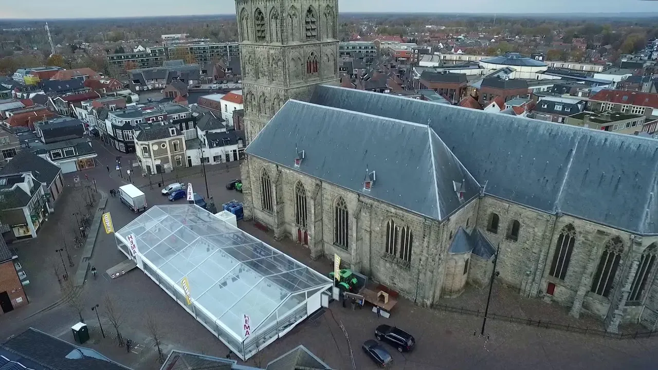 A drone view over Plechelmusplein in Oldenzaal showing the long transparent marquee of the ice rink standing beside the stone Plechelmus basilica, with the old town's roofs around. — IJsbaan Oldenzaal