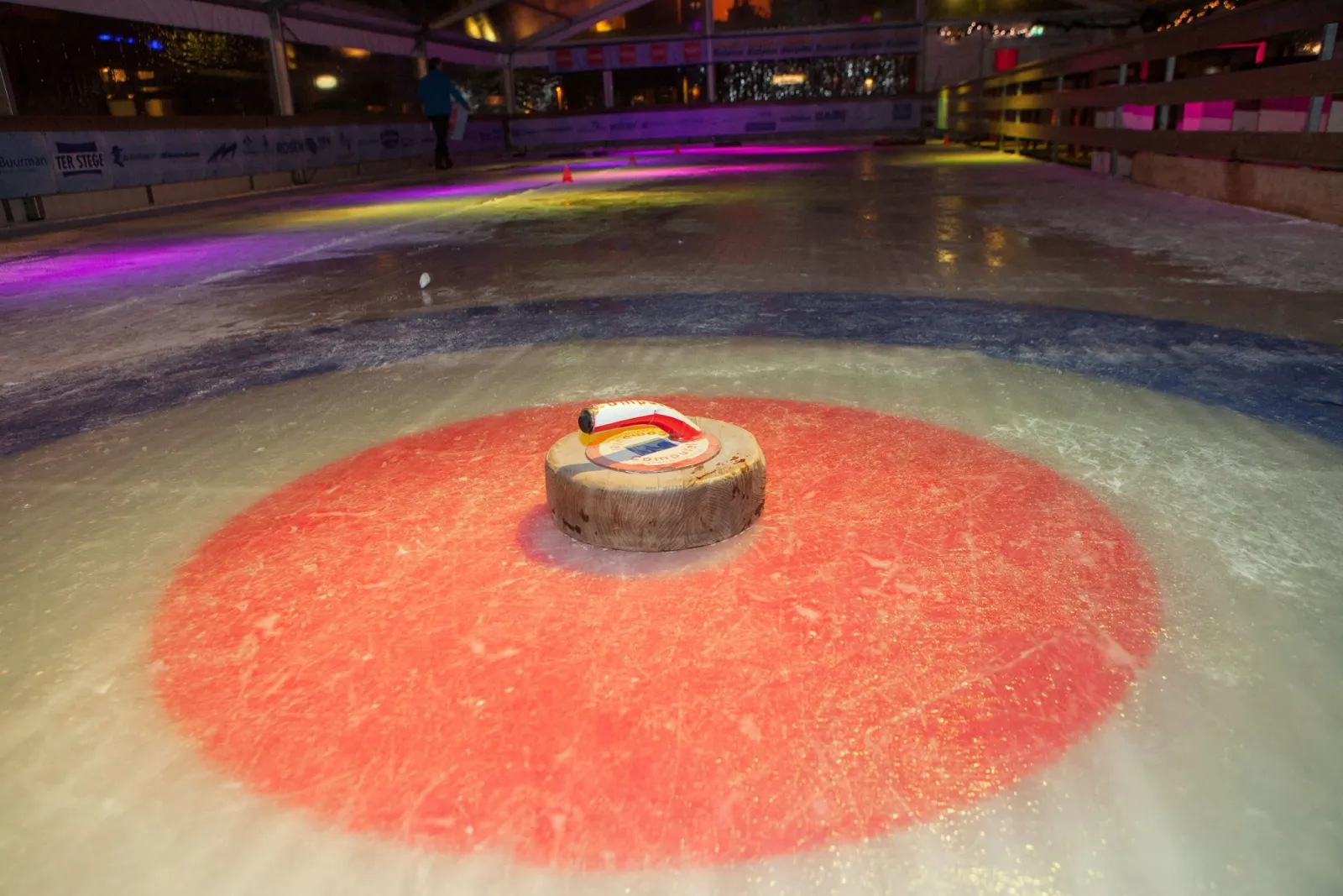 The ice inside the rink's marquee in the evening, with a red curling target and a curling stone in the foreground, coloured lights along the boards and the market stalls behind. — IJsbaan Oldenzaal