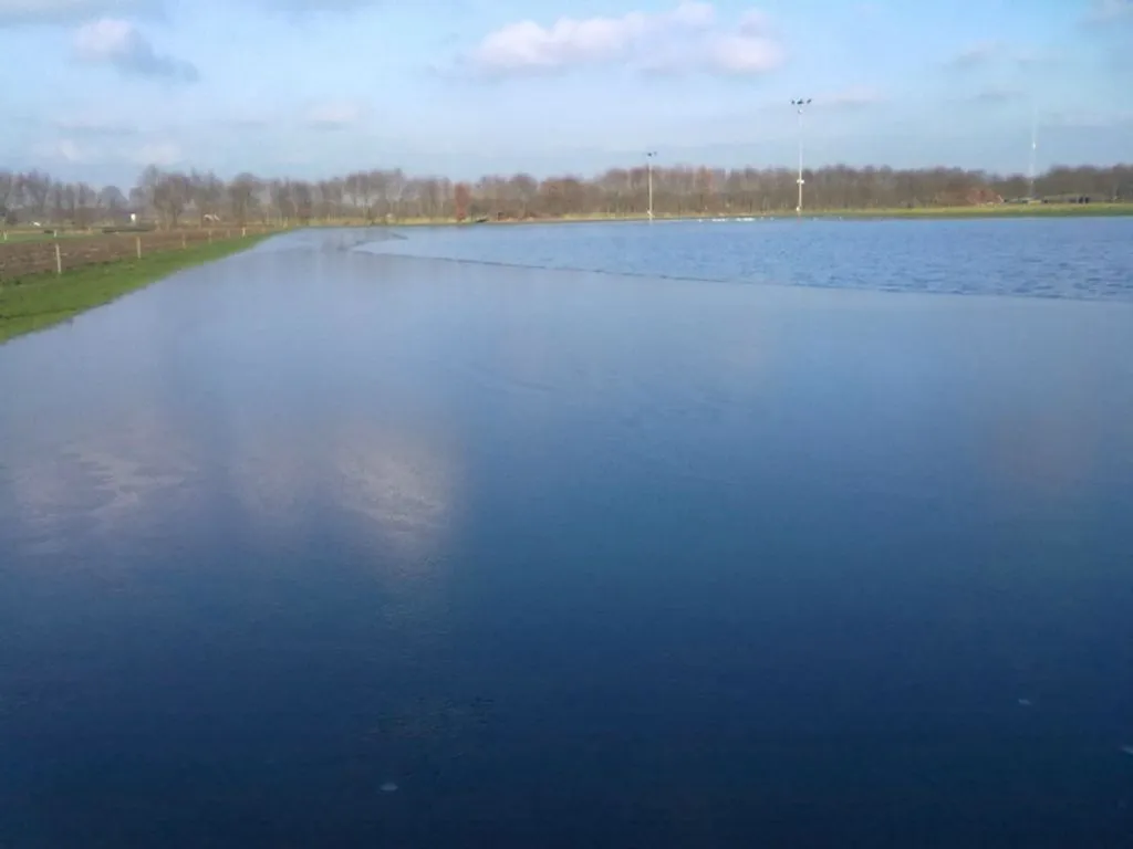 The flooded Ruurlo rink field under a blue sky with floodlight masts along the far edge. — IJsbaan Ruurlo