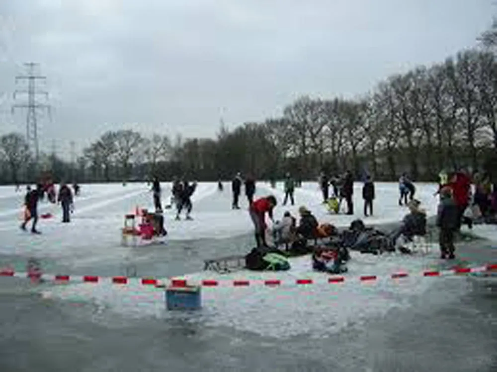 Skaters and families spread over the marked-off ice of the Sietsenveen rink on an overcast day, with a pylon and tall bare trees behind. — IJsbaan Sietsenveen