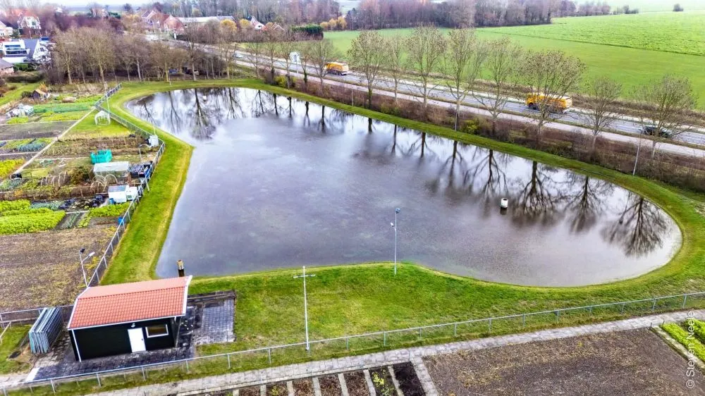 Een dronebeeld van de onder water gezette baan bij 't Winterhof in december 2025, met het zwarte clubgebouw naast het water en velden en een weg erachter. – IJsbaan 't Winterhof