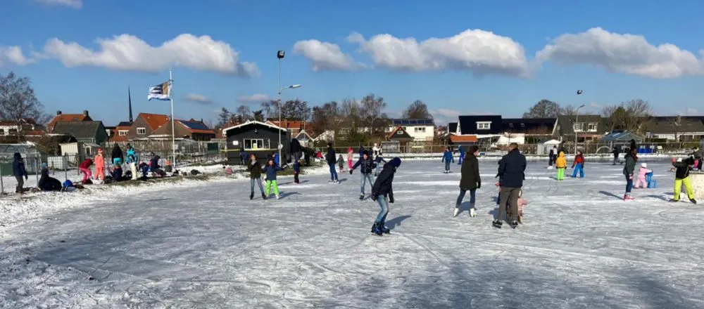 Schaatsers en gezinnen verspreid over het open ijs bij 't Winterhof op een zonnige dag in februari 2021, met huizen en een vlag achter de baan. – IJsbaan 't Winterhof