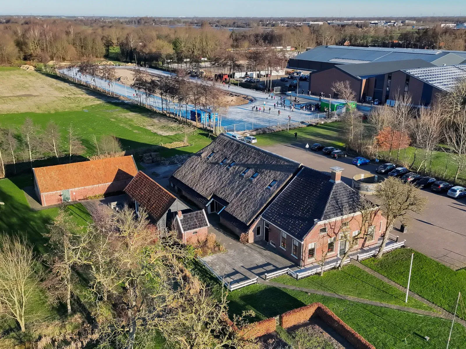 Een dronebeeld van de nieuwe Tolberter baan als een bleek lint van ijs naast de Cazemierboerderij en het bijbehorende parkeerterrein. – IJsbaan Tolbert