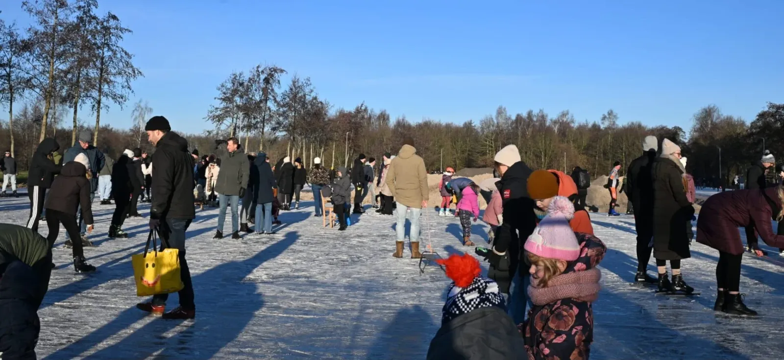 Een groep schaatsers, grotendeels van achteren gezien, op het zonovergoten ijs van de Tolberter baan onder hoge kale bomen. – IJsbaan Tolbert