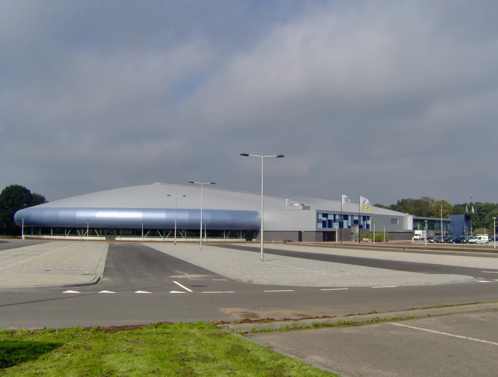 IJsbaan Twente in Enschede seen across its car park, a long low silver and blue domed hall with flagpoles at the entrance end — IJsbaan Twente
