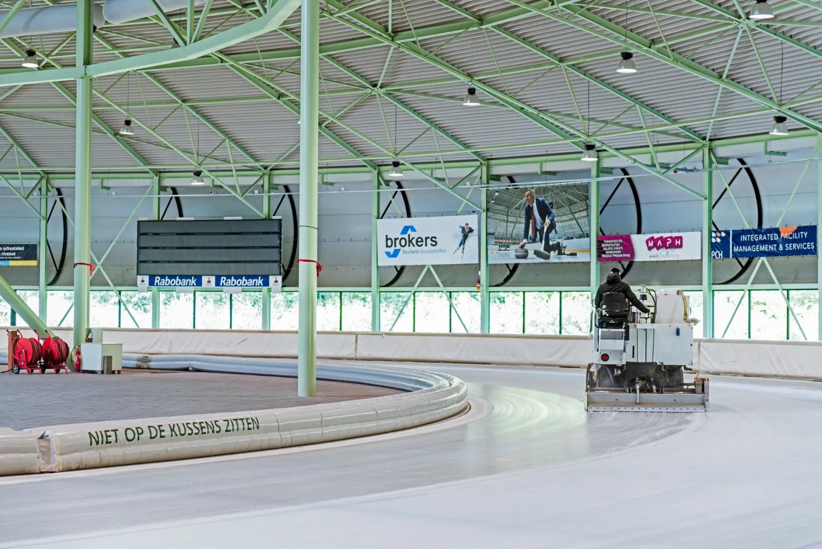 The empty ice of the covered 400 m track at IJsbaan Twente with the ice resurfacer working a bend, under the green steel roof, the boards and advertising panels along the track — IJsbaan Twente
