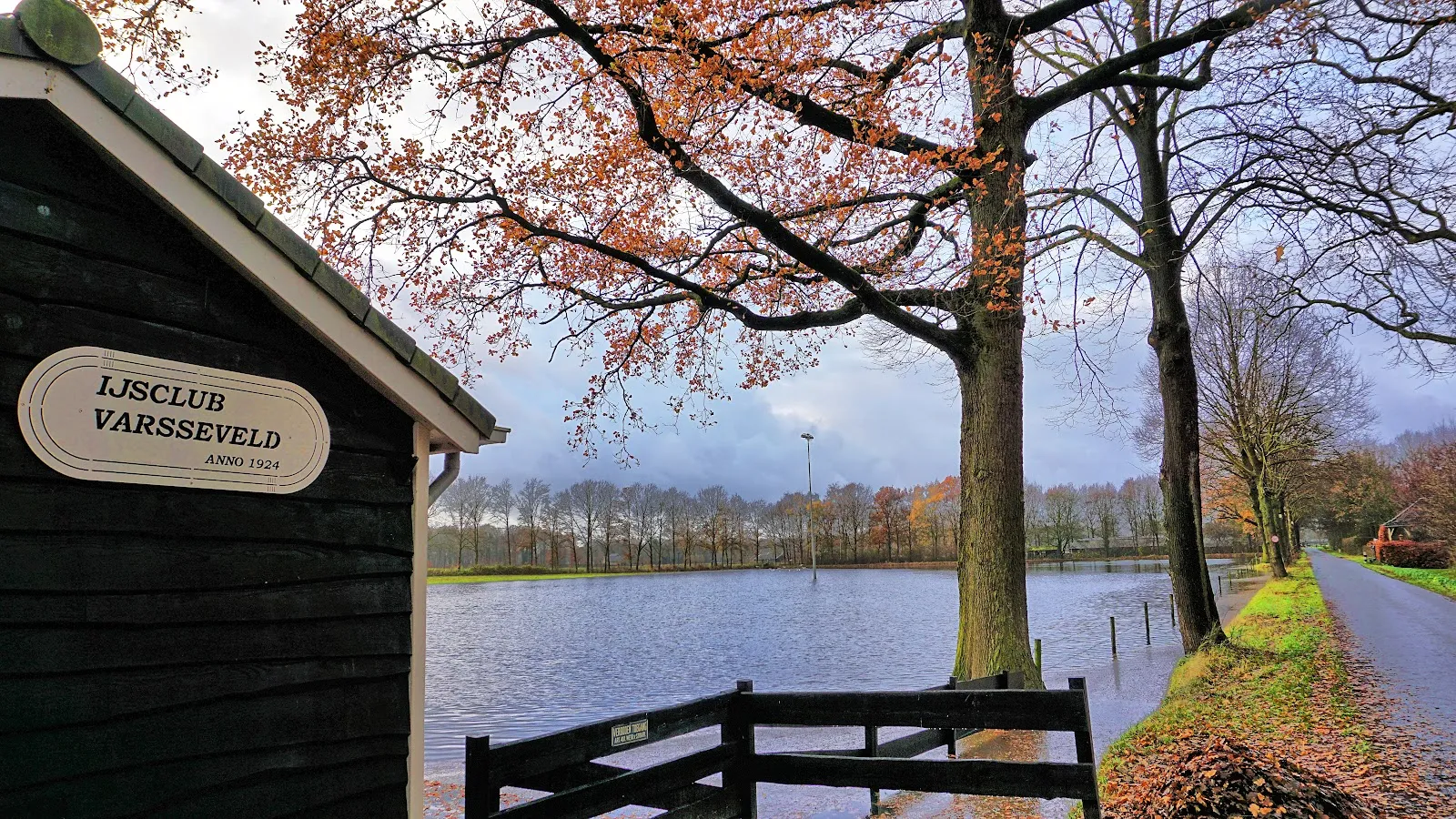 The dark timber clubhouse with its IJsclub Varsseveld sign beside the flooded rink field, framed by autumn trees. — IJsbaan IJsclub Varsseveld