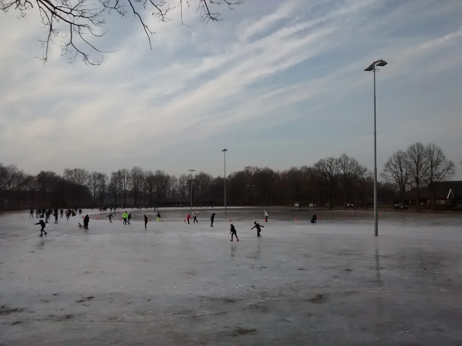 Skaters at the far side of the open natural-ice field of the Varsseveld rink, with floodlight masts and bare trees under a pale sky. — IJsbaan IJsclub Varsseveld