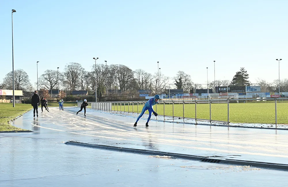 Skaters at a distance on the smooth ice of the Veenoord track on a clear day, with floodlight masts and the sports park behind. — IJsbaan Veenoord