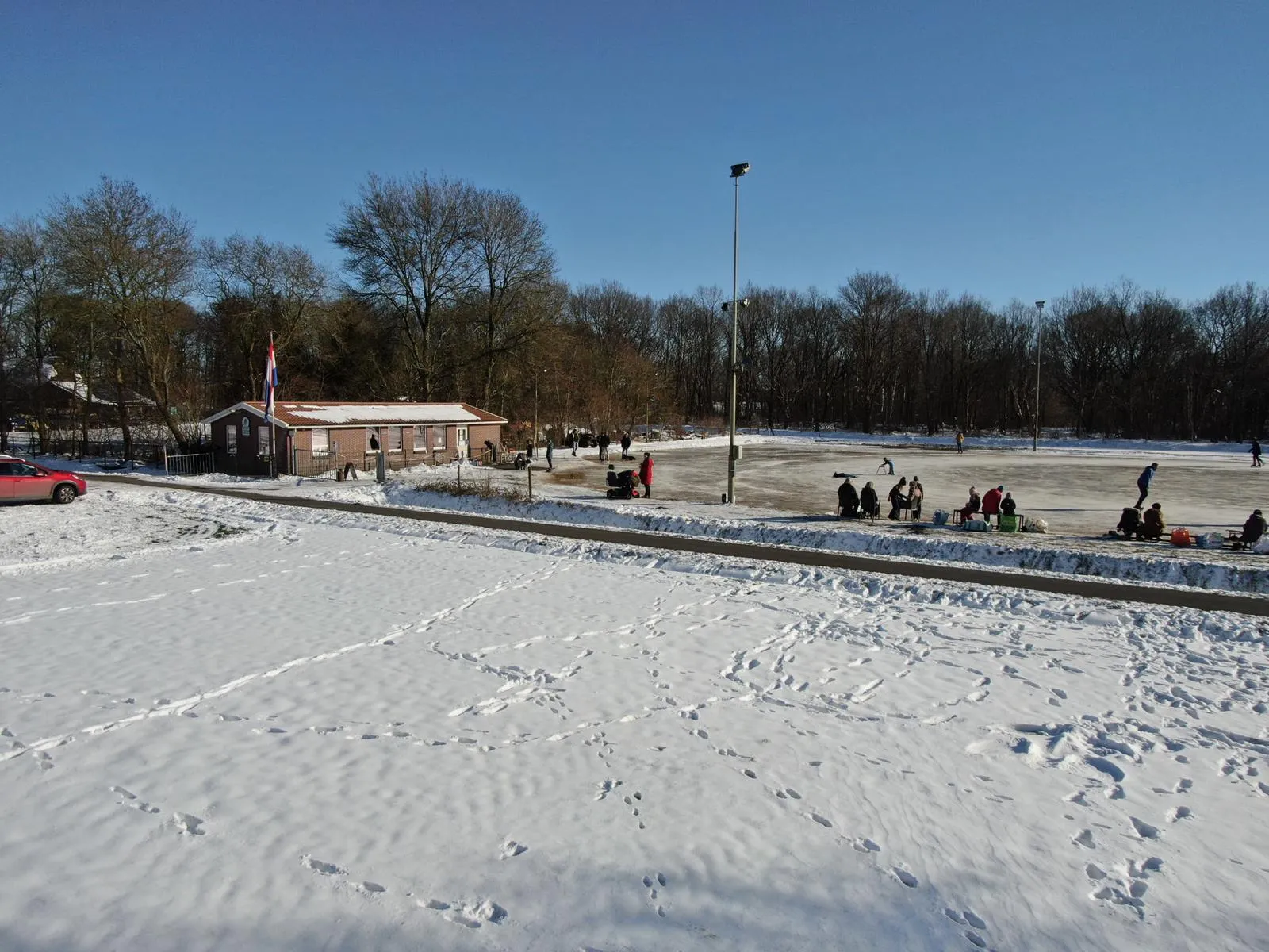 Snow-covered Witten rink with the low brown clubhouse, bare trees and floodlight masts beyond. — IJsbaan Witten