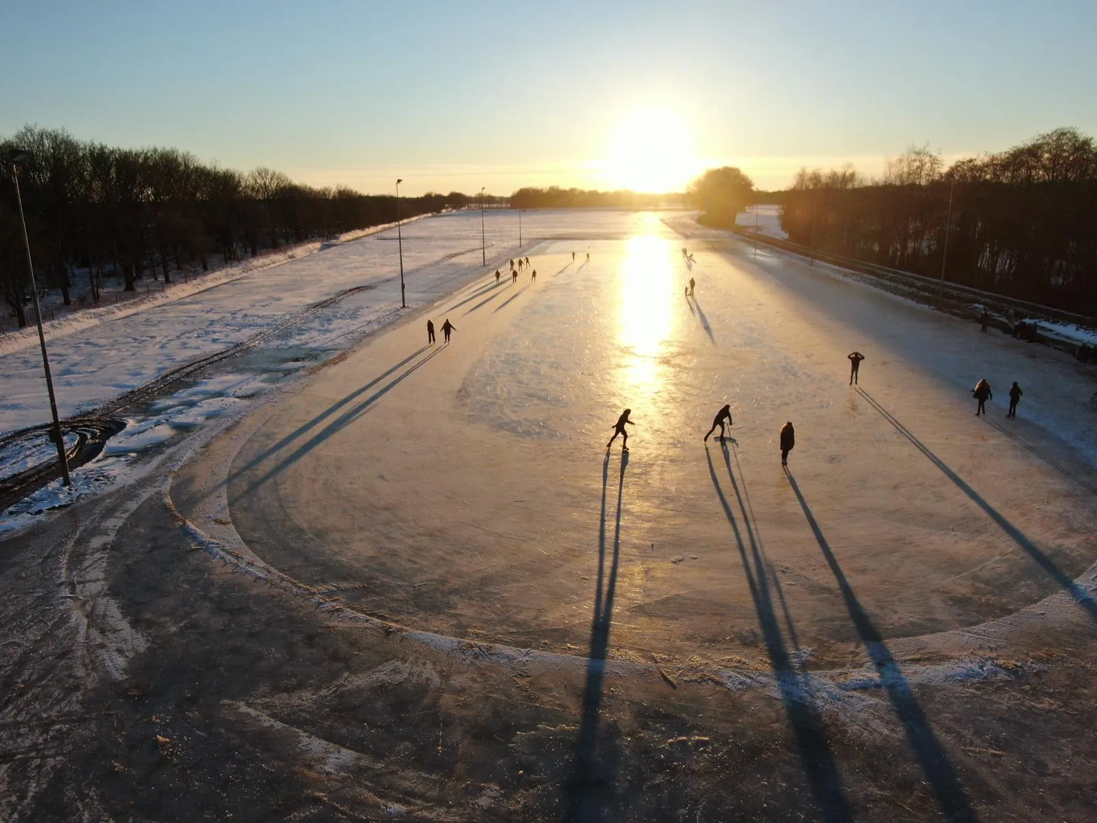 Skaters with long low-sun shadows gliding across the wide Witten rink at sunset, floodlight masts along the edge. — IJsbaan Witten