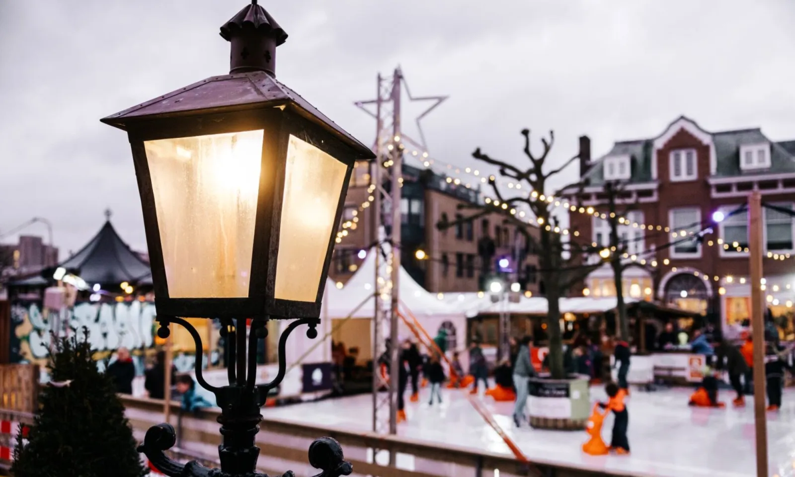 The temporary rink on the Raadhuisplein behind a street lamp, with skaters on the ice and festive lights over the town-centre square. — IJsbaan Zandvoort