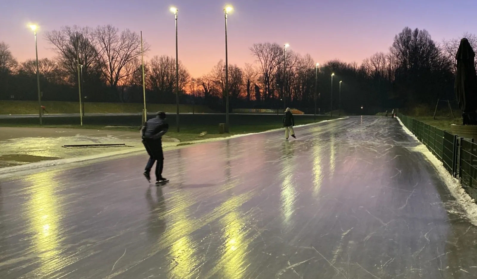 Een lange strook glad natuurijs in Zeist in de schemering, verlicht door hoge lantaarnpalen, met een schaatser op het ijs en kale bomen tegen een roze lucht erachter. – IJsbaan Zeist (Blikkenburg)