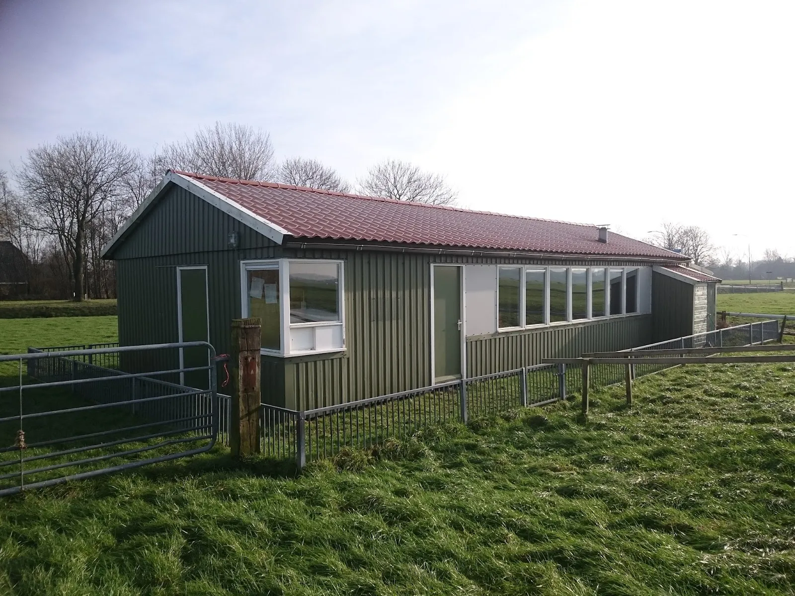 The green timber clubhouse of the Zuidbroek ice rink beside the grass, with a fence around it. — IJsbaan Zuidbroek