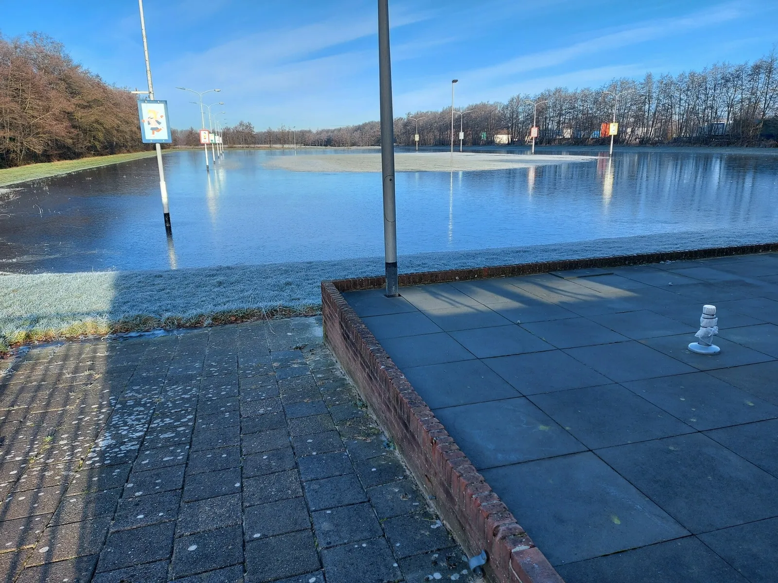 The flooded ice field of the Akkerwoude ice club on a bright winter day: a wide sheet of ice behind a paved edge, with a post and a sign at the front. — IJsbaan Akkerwoude (IJsclub Akkerwoude, De Snûter)