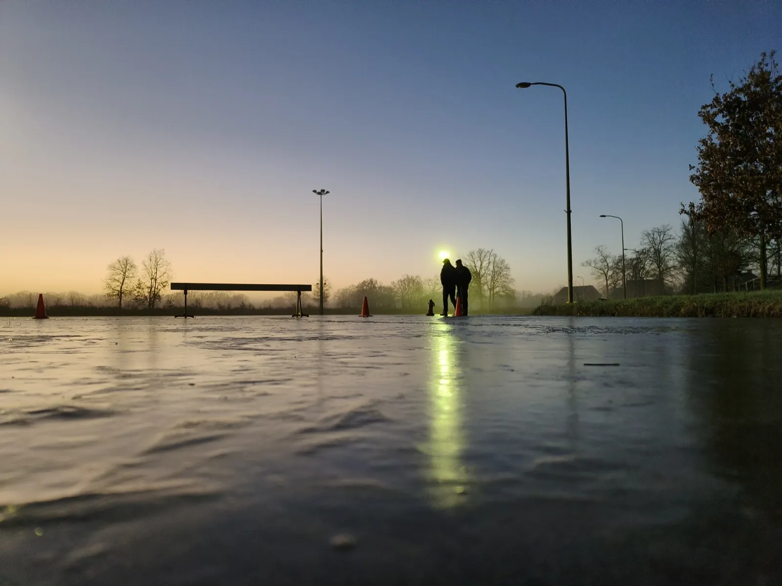 The smooth natural-ice surface at Albergen at dusk, photographed low over the ice, with floodlights reflected in it, a bench and two distant skaters against the sky. — IJsbaan Albergen (IJsclub Albergen)