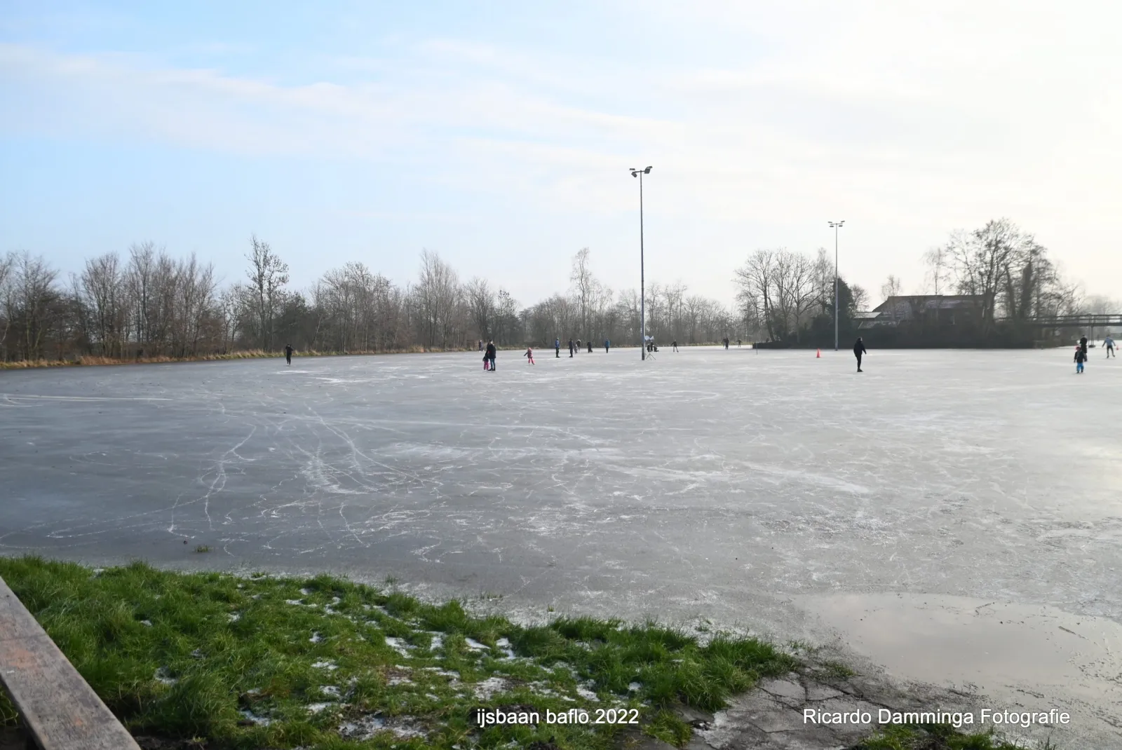 Wide view across the frozen 't Olhof rink in Rasquert with distant skaters, floodlight masts and trees along the far edge. — IJsclub Baflo