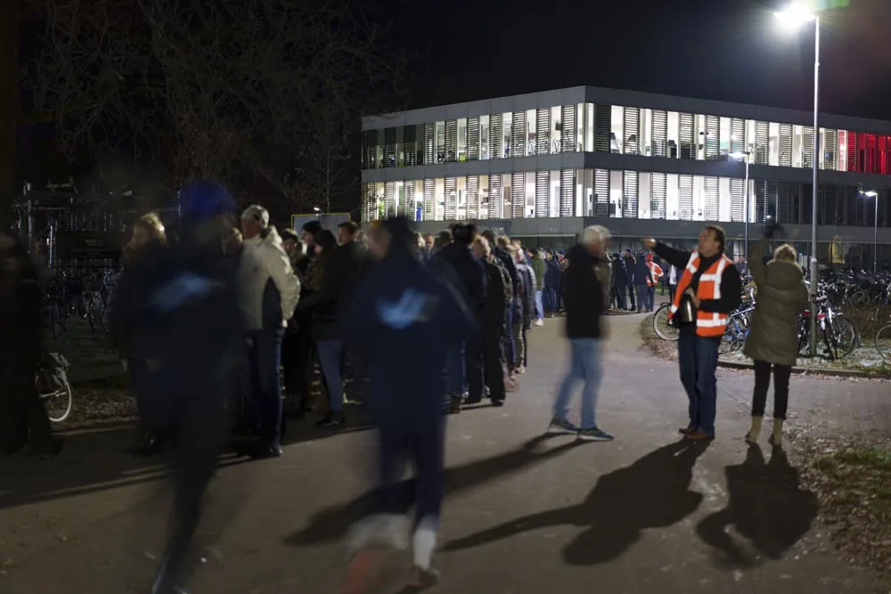 Het verlichte ijsbaangebouw van IJsclub Bergum met twee verdiepingen, 's nachts, met een menigte toeschouwers ervoor en een lichtmast ernaast, tijdens een marathon in 2022. – IJsbaan Burgum (IJsclub Bergum)