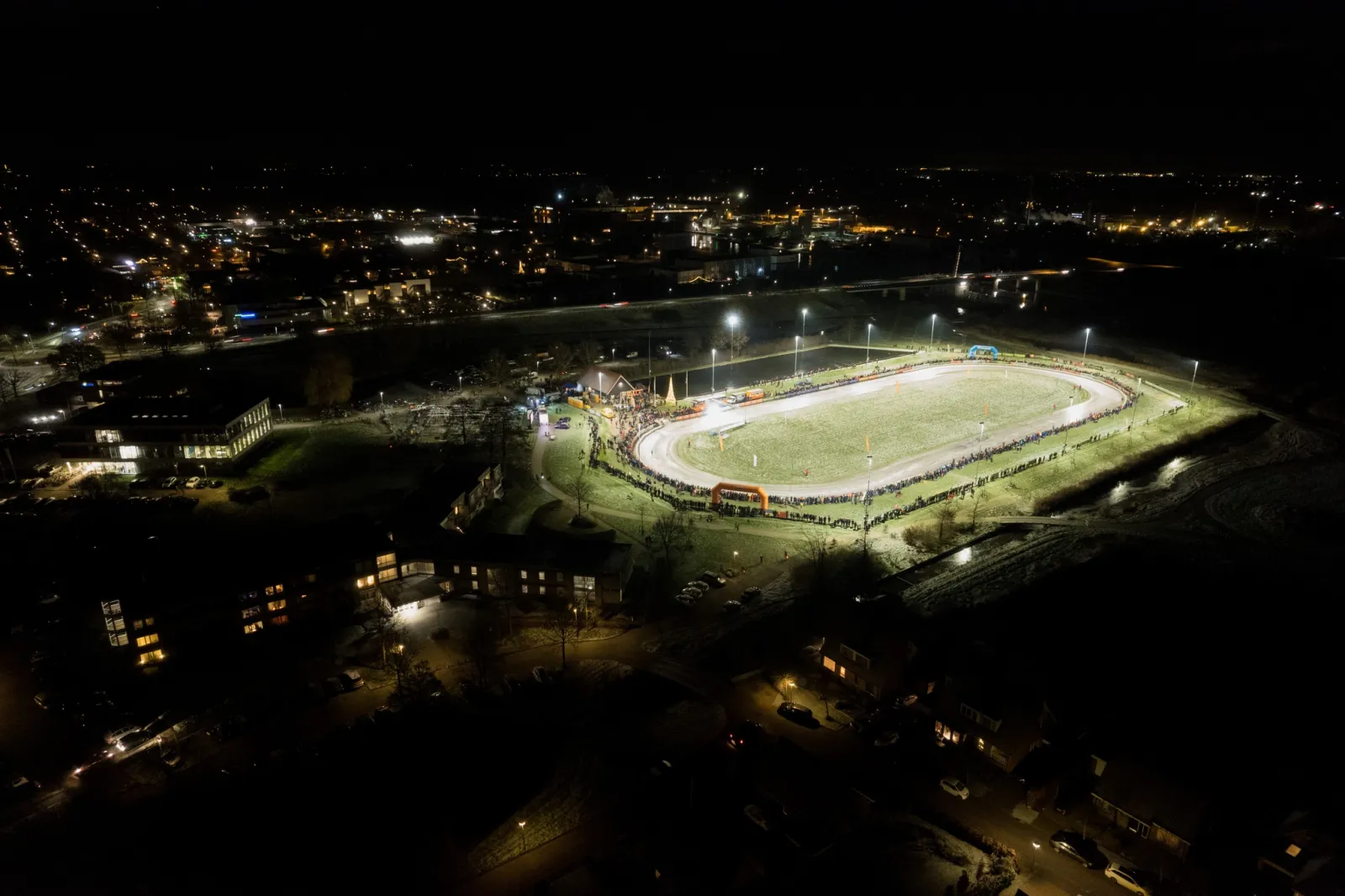 Een luchtfoto in de nacht van de verlichte natuurijsbaan van Burgum, een helderwitte ovaal van ijs met een grasveld in het midden aan de rand van de verlichte straten van het dorp. – IJsbaan Burgum (IJsclub Bergum)