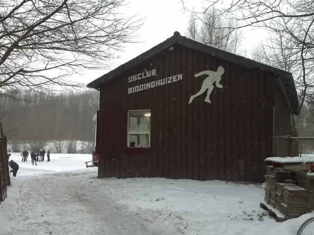 Het donkerrode houten clubgebouw van IJsclub Biddinghuizen met de clubnaam en een schaatserfiguur op de gevel, in de sneeuw, met schaatsers en kale bomen erachter. – IJsbaan Biddinghuizen (IJsclub Biddinghuizen)