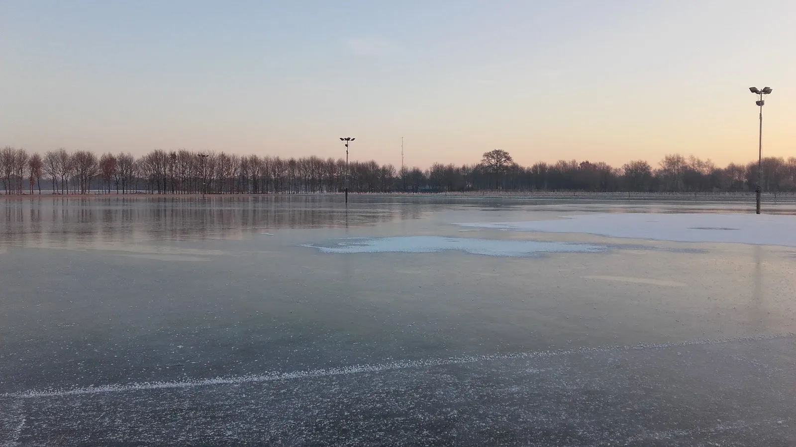 De bevroren baan van IJsclub Borculo in de schemering, met een lichtmast en een rij bomen langs de overkant van het ijs. – IJsclub Borculo