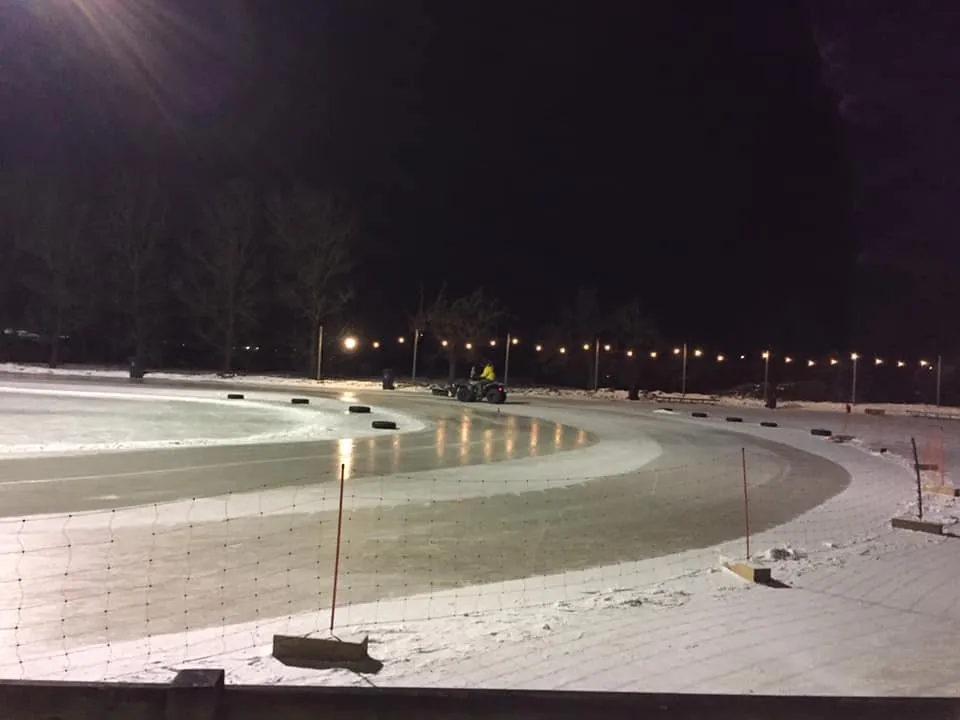 De verlichte bocht van de hoofdbaan 's avonds met lichtslingers aan de verre kant, een quad met een ijsonderhoudsaanhanger en een net hek op de voorgrond. – IJsclub Centrum Lopik