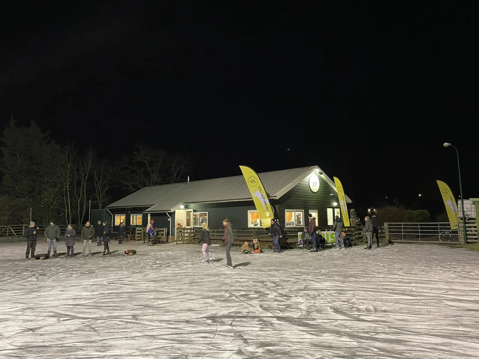 Schaatsers en gezinnen op het verlichte ijs 's avonds voor het donkergroene clubgebouw van Lopik, met gele vlaggen en kale bomen erachter. – IJsclub Centrum Lopik