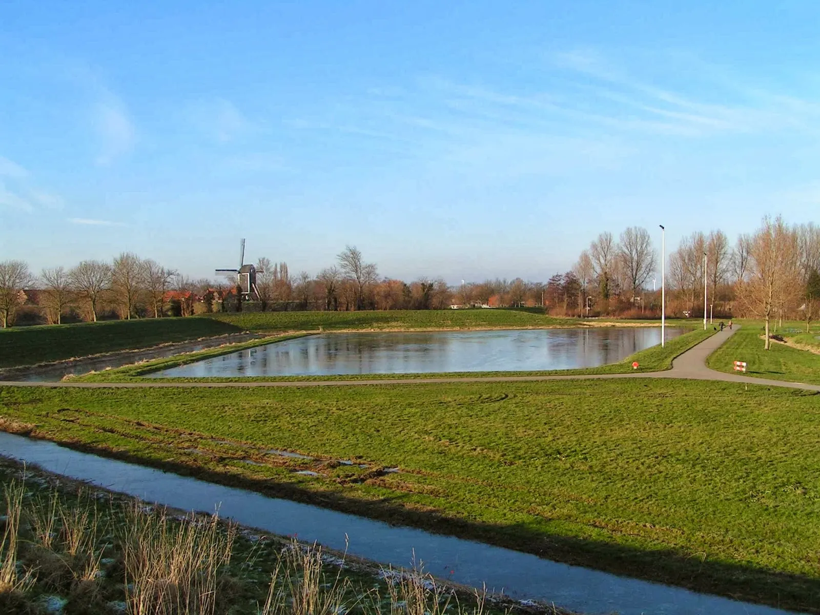 The Brielle rink basin beside the cycle path under a blue sky, with a windmill and floodlight masts beyond. — IJsclub De Brielsche Geuzen