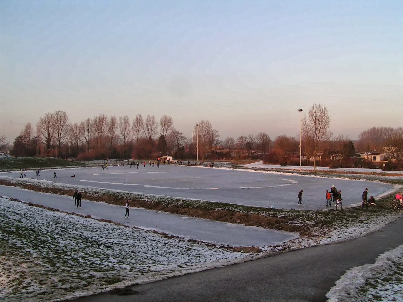 Skaters spread across the frozen Brielle rink basin at sunset, with floodlight masts and bare trees along the banks. — IJsclub De Brielsche Geuzen