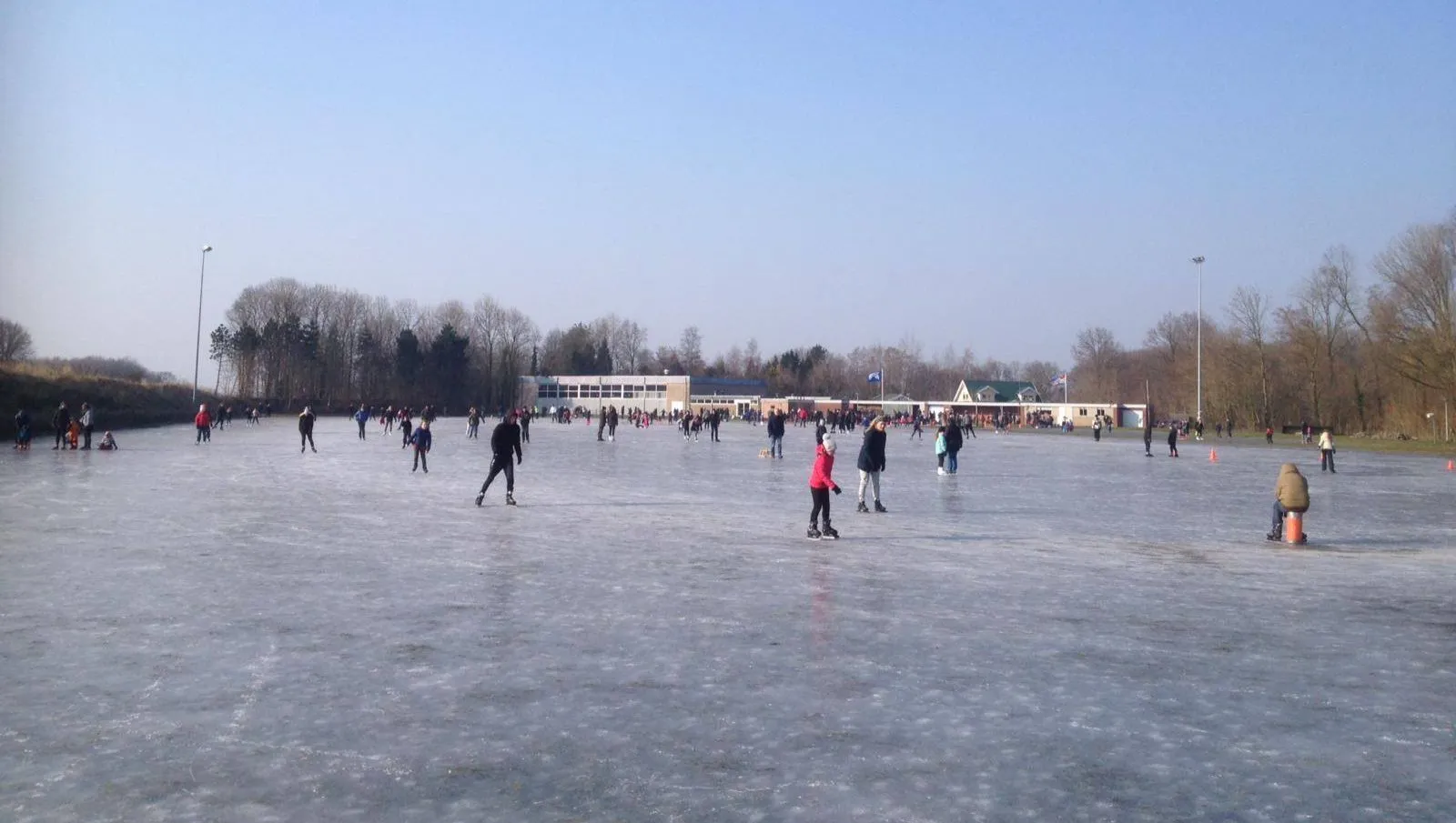 Skaters at a distance across the wide natural ice of the Oosterwolde rink under a clear blue sky, with the long low clubhouse and bare trees along the far side. — IJsbaan De Slatten (IJsclub De Eendracht, Oosterwolde)