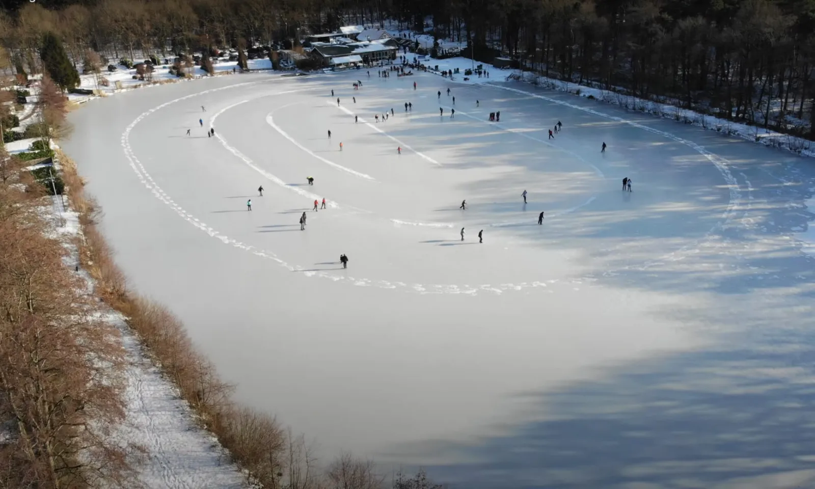 Luchtfoto van de bevroren baan van de Falieberg met schaatsers in de verte en het clubgebouw naast het ijs. – IJsclub de Falieberg