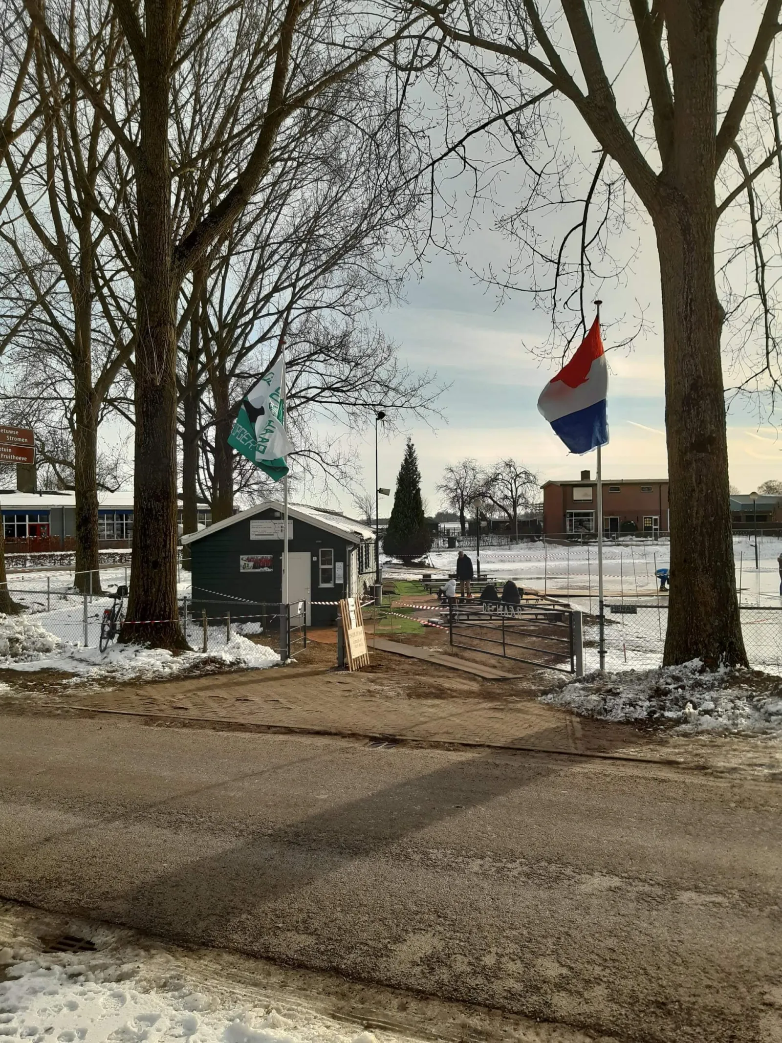 The green clubhouse hut at the rink entrance on Korte Veersteeg, with the club flag and a Dutch flag, picnic benches beside the ice fence and a few visitors sitting in the distance. — IJsbaan De Haar (Enspijk)