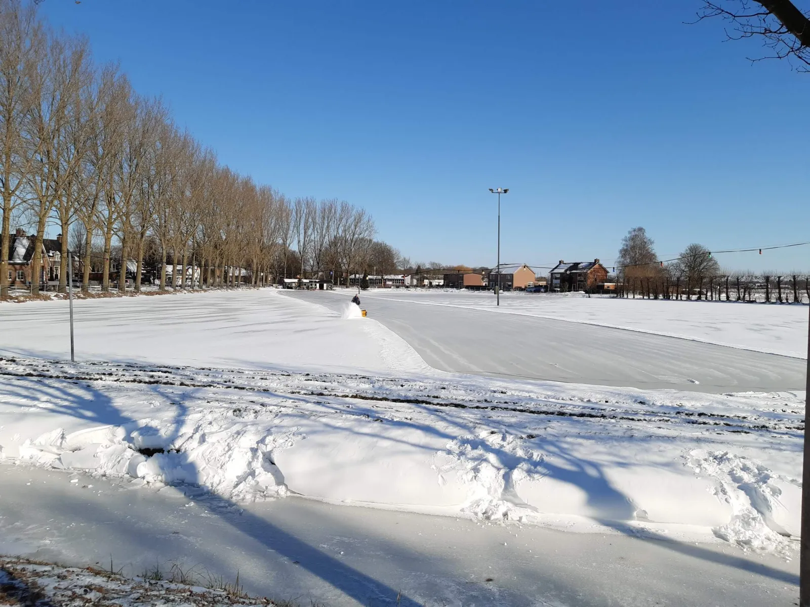 The snow-swept ice field at Enspijk in February 2021, with a volunteer clearing a lane with a snow blower, a floodlight mast, a row of poplars and village houses behind. — IJsbaan De Haar (Enspijk)