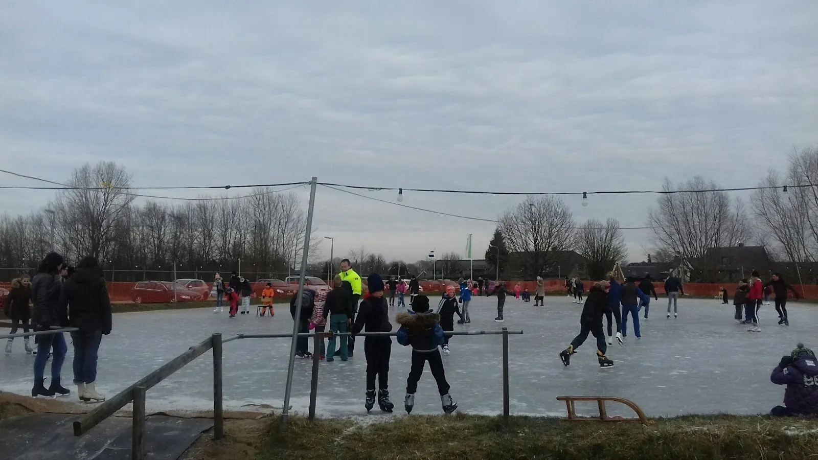 Schaatsers en kinderen op de omrande natuurijsbaan van IJzendoorn op een grauwe winterse middag, met lichtslingers boven het ijs en kale bomen erachter. – IJsclub De Hoeken