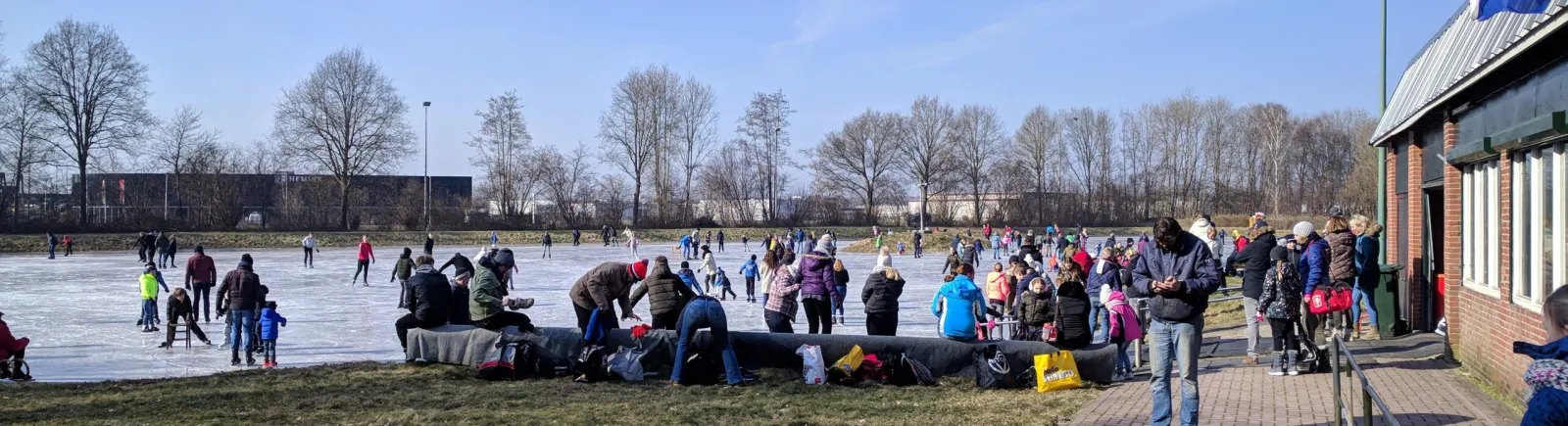 Skaters and families on the open natural ice at Holten beside the red-brick clubhouse on a bright winter day. — IJsclub De Noordpool Holten